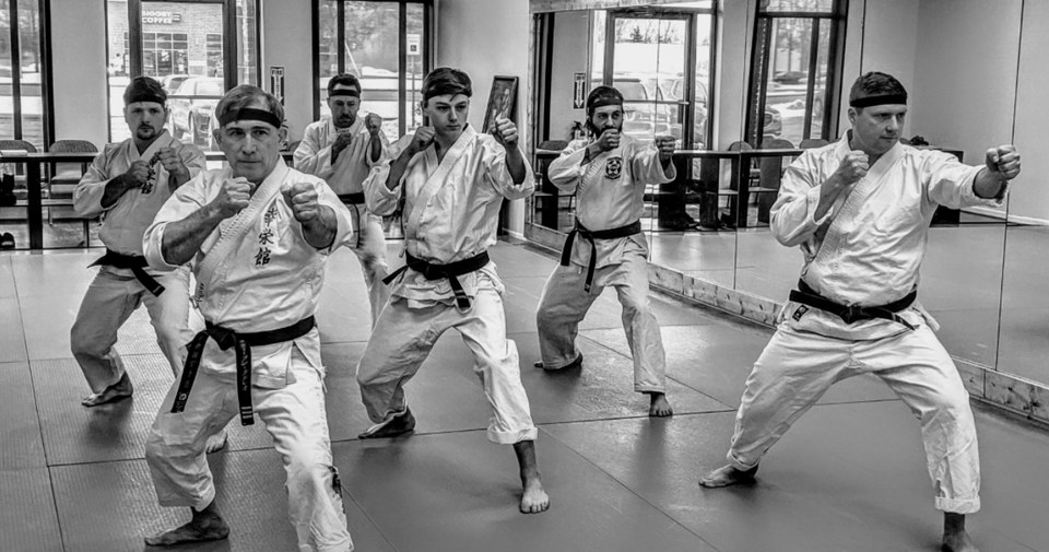 Six black belts in white gis practicing a stance together on the dojo mats